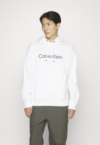 Witte katoenen hoodie met een voorzak, capuchon met trekkoord en het zwarte 'Calvin Klein' logo. Gepresenteerd op een model in olijfgroene cargobroek.