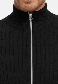 Svart stickad tröja med hög krage, med ett texturerat kabelmönster och en silverfärgad dragkedja som löper ned längs framsidan.