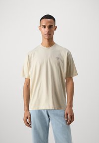 Tricou din bumbac de culoare crem, cu mâneci scurte, decolteu rotund și un mic patch cu logo triunghiular pe pieptul stâng.