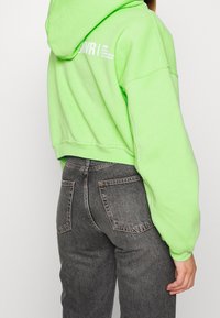 Felpa corta con cappuccio verde lime, con scritte bianche sulla schiena e maniche lunghe. Abbinata a jeans grigi a vita alta.