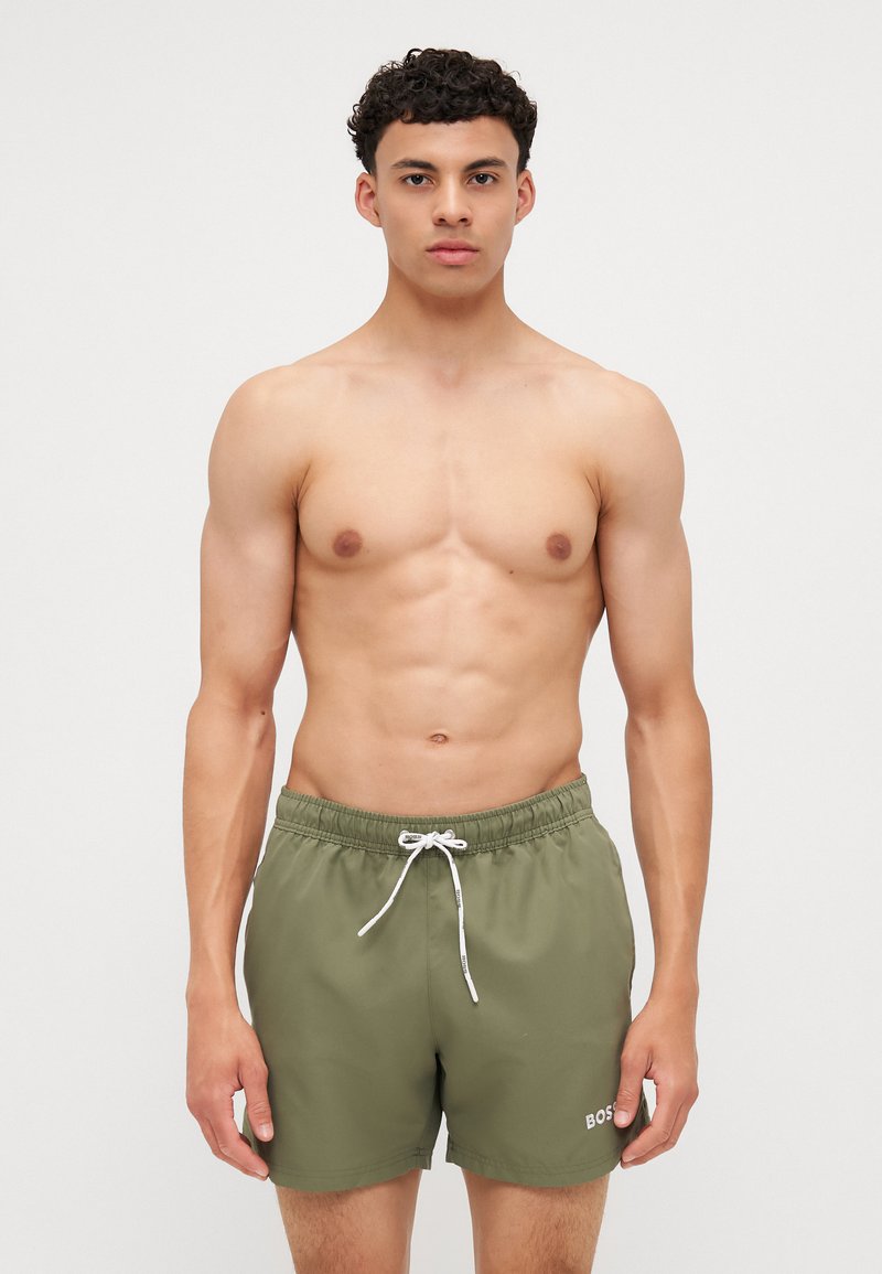 Short de bain vert olive avec une taille élastique et un cordon blanc. Présente un petit logo en bas à droite. Texture lisse.