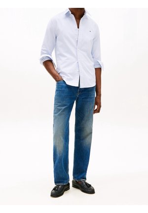 Mand iført lysblå stribet skjorte med knapper, blå jeans med lige ben og sorte loafer med kvaster, står med den ene hånd i lommen.