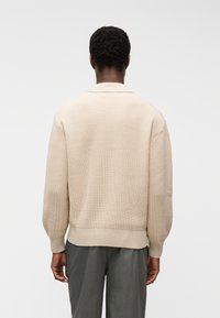 Άνδρας με σκούρο δέρμ α που φοράει μπεζ πλεκτό πουλόβερ με ρίγες και σκούρο γκρι παντελόνι, εμφανιζόμενος από πίσω με φόντο έναν απλό φωτεινό τοίχο.