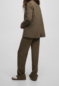 Brown tailored blazer with a relaxed fit, single back vent, and matching loose pants. Worn with white sneakers featuring brown accents.