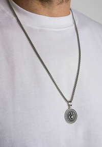 Colier din argint cu un pandantiv circular în formă de leu, cu detalii intricate și o margine texturată, purtat peste un tricou alb.