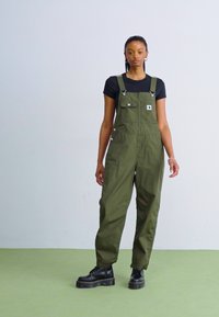 Olive green overalls with adjustable shoulder straps, front pockets, and a zippered opening, paired with a fitted black shirt and black boots.