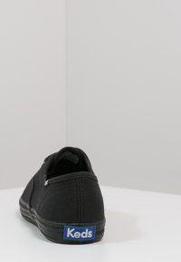 Musta kangastenniskenkä pyöreällä muodolla, tasaisella pohjalla ja sinisellä Keds-logolla takana. Ominaisuuksiin kuuluu minimaalinen ompelu ja sileä pinta.