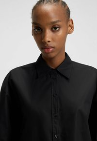 Donna con capelli intrecciati che indossa una camicia nera con bottoni, in un ambiente chiaro e uniforme, rivolta in avanti con un'espressione neutra.