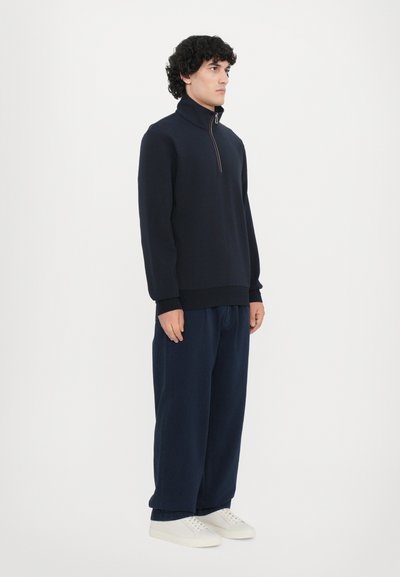 Sudadera con cremallera en navy de tejido suave, con un alto cuello y puños acanalados. Combinada con pantalones anchos en navy y zapatillas blancas.