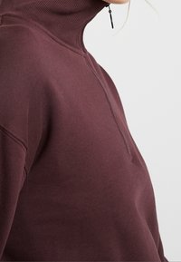 Primer plano de una persona que lleva un suéter de cuello alto burdeos con media cremallera y un tirador de cremallera negro, contra un fondo liso.