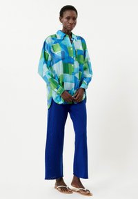 Leichte, langärmlige Bluse in blau-grünen geometrischen Mustern, kombiniert mit strukturierten blauen weiten Hosen und lässigen Sandalen.