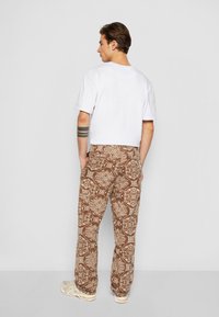 Hvit kortermet t-skjorte kombinert med brune mønstrede bukser med intrikate design. Modellen står sideveis, iført lyse joggesko.