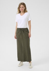 T-shirt blanc à manches courtes et décolleté en V associé à une longue jupe olive avec taille à cordon de serrage et deux poches avant. Porté avec des baskets beiges.