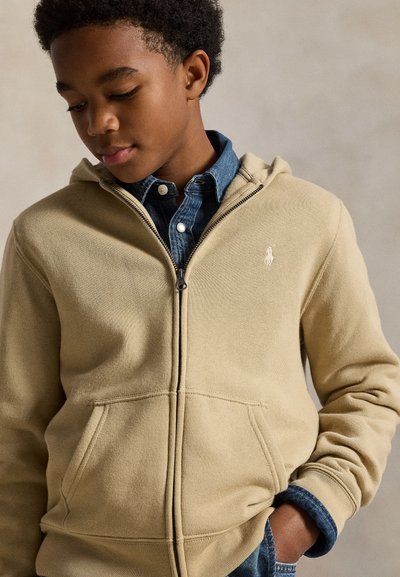 Jongen draagt een beige zip-up hoodie over een blauwe spijkerblouse met knopen, kijkt naar beneden met één hand in de zak tegen een effen achtergrond.