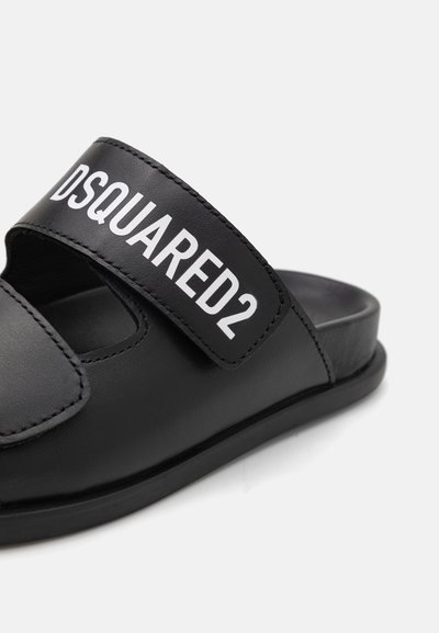 Musta liukusandaali, jossa on leveä brändätty hihna, jossa lukee "DSQUARED2" valkoisella, valmistettu sileästä synteettisestä materiaalista ja jossa on litteä pohja.