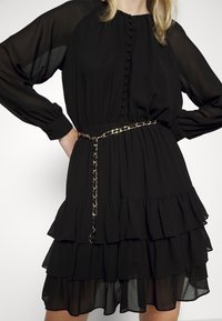 Femme portant une robe noire à plusieurs couches avec des manches longues transparentes, des poignets boutonnés, un devant boutonné et une ceinture chaîne dorée à la taille.