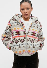Felpa con zip in crema, morbida e pelosa, decorata con motivi tribali multicolore. Presenta un cappuccio, due tasche frontali e polsini e orlo a costine.