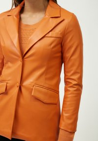 Blazer de piel sintética naranja con solapas de muesca, dos bolsillos frontales y cierre de botón, combinado con un suéter texturizado a juego debajo.