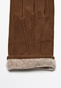 Bruine suède handschoen met een gebreide beige manchet, voorzien van subtiele stikdetails op de achterkant voor textuur en visuele belangstelling.