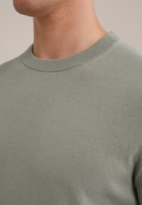 Ljusgrön tröja med rundad halsringning i mjukt, texturerat tyg, med en ribbad halsringning och slät yta. Nära vy.