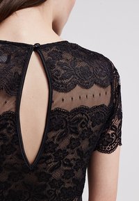 Svart spetstopp med korta ärmar, med blommönster, en nyckelhålsöppning på ryggen och en knappstängning vid halsringningen. Genomskinlig textur överlag.