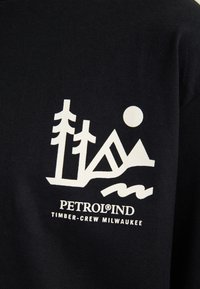 Mustassa kankaassa valkoinen graafinen kuvasto, jossa on abstrakteja vuoria, puita ja aurinko, ja teksti "PETROLIND TIMBER-CREW MILWAUKEE."