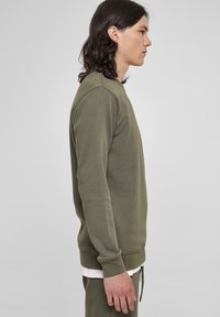 Olijfgroen sweatshirt met een ronde hals, ribgebreide manchetten en zoom. Zacht, gestructureerd materiaal van opzij getoond met een gelaagd wit shirt eronder.