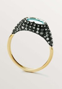 Anillo en tono dorado que presenta una gran piedra azul, rodeada de piedras negras y transparentes, con un diseño ondulado y texturizado en la banda.