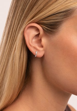 Arete de plata en forma de aro con un diseño texturizado, que presenta una forma curva y un acabado suave, llevado en la oreja izquierda. Estilo minimalista.