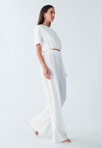 Biały dwuczęściowy strój składający się z krótkiego, teksturowanego topu z krótkimi rękawami oraz wysokich, szerokich spodni. Regulowany pas z wiązaniem ozdobionym koralikami.