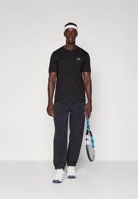 Svart sporttshirt med korta ärmar och en logotyp, kombinerad med mörka byxor. Håller i ett tennisracket, bär vita sneakers och ett vitt pannband.