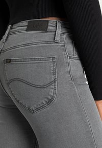 Acercamiento a una persona que lleva puestos unos pantalones Lee grises y un suéter negro de canalé, destacando los detalles del bolsillo trasero y la cinturilla.