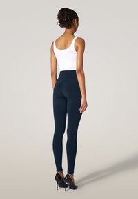 Marina leggings med hög midja, slät tygstruktur och en åtsittande design, kombinerat med en vit ärmlös topp och svarta klackar.