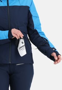 Marineblau und blaue Skijacke mit einer Reißverschlusstasche, ausgestattet mit einem Logopatch und elastischen Bündchen. Glattes Material mit angepasstem Schnitt.
