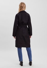 Cappotto lungo nero con vita regolabile, dotato di colletto, maniche lunghe e taglio dritto. Indossato sopra dei jeans blu e stivali neri con tacco.