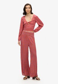 Conjunto de dos piezas con patrones en rojo y rosa, que incluye una blusa de manga larga con un corte en forma de ojal y pantalones de pierna ancha de cintura alta. Textura suave.