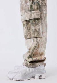 Camo-gepatroneerde cargo-broeken in beige en groene tinten, gecombineerd met witte en zilveren Nike sneakers die zijn voorzien van opvallende gelaagde demping.