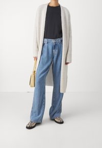 Světlý šedý dlouhý kardigan přes černé tričko, doplněný modrými širokými džíny a botami s hadím vzorem. Drží malou žlutou kabelku.