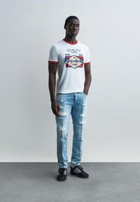 Jeune homme debout, une main dans la poche, portant un t-shirt blanc à motif avec des bordures rouges, un jean bleu clair déchiré et des baskets noires à rayures blanches.