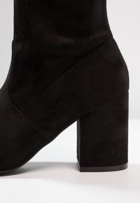 Botín negro de ante hasta el tobillo con un tacón ancho y cuadrado. Presenta una textura suave y un diseño anguloso, con costuras delineadas a lo largo de la costura.