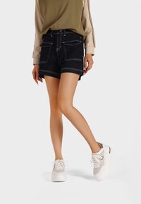 Shorts in denim blu scuro con cuciture bianche, dotati di tasche anteriori e posteriori. Abbinati a sneakers bianche e metalliche con dettagli testurizzati.