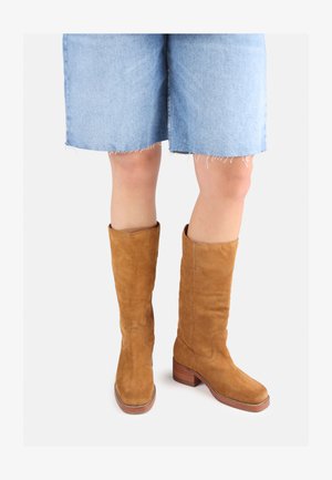 Brune suedestøvler knæhøje med blokhæle og syninger, parret med denimshorts med frynset kant.