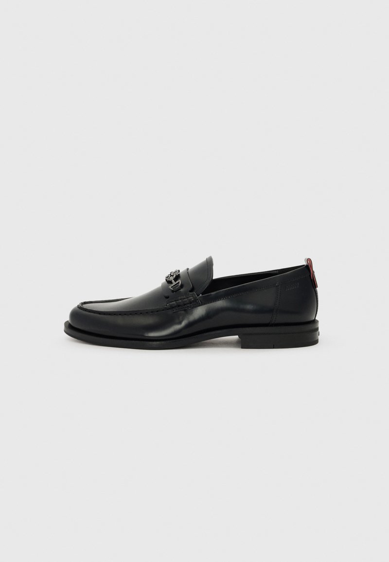 Sorte skinnloafers har en rund tå, hevet sømdetalj, et dekorativt metallakzent på forsiden, og en flat såle med minimal mønstring.