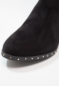 Bota de ante negra con puntera puntiaguda y suela plana, adornada con tachuelas metálicas a lo largo de la base. Textura suave con un acabado liso.