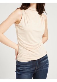 Mujer vestida con una blusa sin mangas de cuello alto color beige y jeans azul oscuro, con las manos apoyadas en la parte baja de la espalda y la cadera.