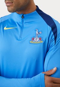 Bl�å langærmet sports top med halv-lynlås ved halsen. Har marineblå detaljer og et logo med en fugl og tekst på brystet. Glat tekstur.