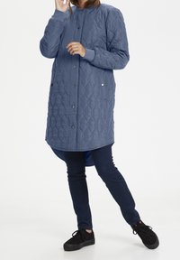 Gepatchter blauer langer Mantel mit Kapuze, Druckknöpfen und seitlichen Taschen; kombiniert mit dunklen Jeans und schwarzen Turnschuhen. Glatte Stoffstruktur.