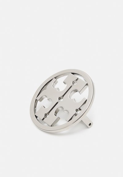 Metallisk ovalt tillbehör med utskuret mönster som har symmetriska former, polerad silverfinish och en fästanordning på sidan.
