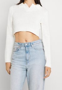 Vit kort tröja med V-ringning och klassisk krage, stickad textur, ribbad nederkant, i kombination med högmidjade ljusblå denimjeans.