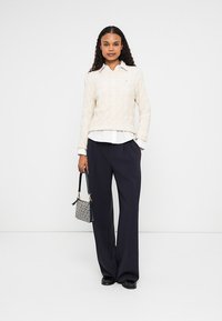 Woman standing wearing cream sweater over white shirt, wide black trousers, black shoes, holding small patterned handbag.
